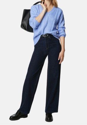 Mujer con suéter de punto trenzado azul claro, vaqueros anchos oscuros con cinturón, mocasines negros, sosteniendo un bolso negro de hombro.