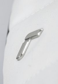 Clip en métal argenté avec une forme épurée, fixé à une surface en tissu blanc texturé. Le clip a une extrémité plate et une légère courbure.