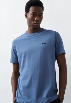 Νεαρός άνδρας με κοντά σγουρά μαλλιά, φορώντας ένα ανοιχτό μπλε T-shirt BOSS, μπροστά σε ένα απλό λευκό φόντο.