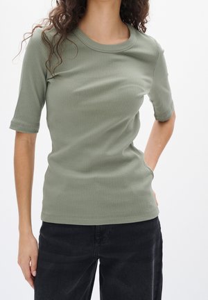 Vrouw draagt een saliegroene geribbelde top met korte mouwen en zwarte broek, staand met één hand in de zak tegen een witte achtergrond.