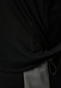 Top atlético negro con un acabado texturizado, que presenta una etiqueta de logo, detalle de cordón y un panel de malla gris en la parte inferior.