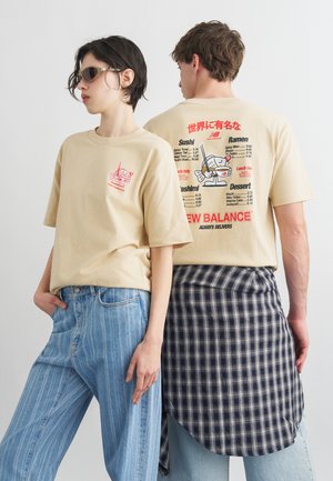 Twee mensen dragen beige New Balance T-shirts met rode sushi-menugraphics; de een kijkt frontaal met een zonnebril, de ander toont de achterkant met een geruit overhemd om de taille gebonden.