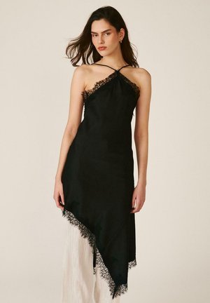 Mujer con vestido lencero asimétrico negro con ribete de encaje y tirantes finos, combinado con pantalones anchos blancos plisados.