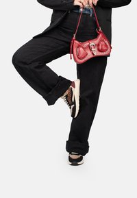 Borsa a tracolla in pelle rossa con decorazioni a forma di cuore e borchie. Indossata con jeans larghi neri e sneaker multicolori con un design a fantasia.