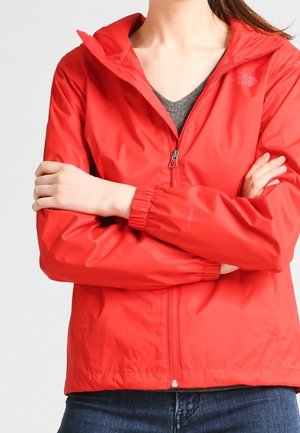 Kvinde iført en rød North Face-jakke med lynlås og oprejst krave, grå skjorte under, krydsede arme og synlige blå jeans.