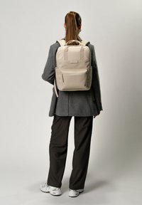 Sac à dos beige avec deux poignées supérieures et des bretelles réglables, comportant une poche zippée à l'avant et une poche en mesh sur le côté, présenté sur une personne.