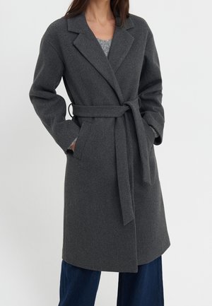 Femme portant un manteau en laine gris foncé ceinturé, mi-long, avec de larges revers et des poches latérales, associé à un pantalon bleu foncé.