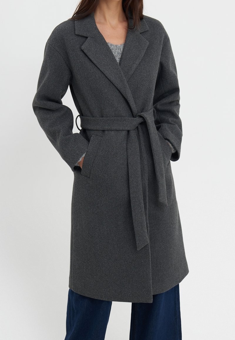 Femme portant un manteau en laine gris foncé ceinturé, mi-long, avec de larges revers et des poches latérales, associé à un pantalon bleu foncé.