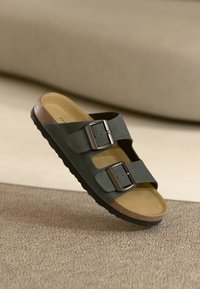 Black suede sandal with two metallic buckles, brown footbed, and textured black sole. Open-toe design with an anatomical shape.