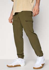 Olive green cargo pants with elastic cuffs and multiple side pockets, paired with white athletic shoes featuring a textured upper and gum sole.