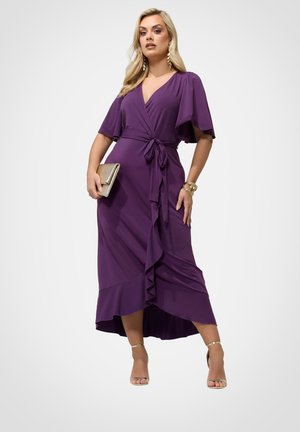 Kvinde iført en lilla slå-om-kjole med flagrende ærmer og flæsekant, der holder en guldfarvet clutch, gyldne armbånd og gyldne højhælede sandaler.