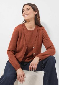 Vrouw met lang, donker haar die een roestkleurige cardigan en trui draagt, donkerblauwe jeans, glimlachend, zittend op een witte, gestructureerde kubus.