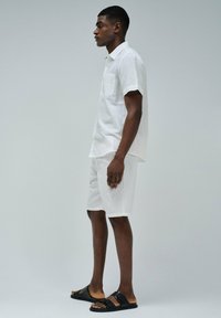 Camisa blanca de lino de manga corta con un bolsillo en el pecho, combinada con pantalones cortos blancos y sandalias negras con dos tiras y suela plana.