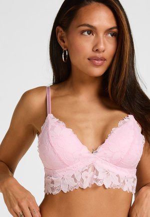 Femme portant un bralette en dentelle rose clair avec des motifs floraux, des boucles d'oreilles anneaux, et de longs cheveux foncés regardant légèrement vers la droite sur un fond uni.