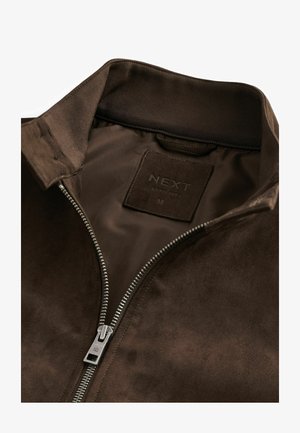 Brūns zamšādas jaciņa ar rāvējslēdzēju priekšā, ribotu apkakli un iespiestu etiķeti ar uzrakstu "NEXT ESTD 1982 M." Gluda tekstūra un minimālistisks dizains.