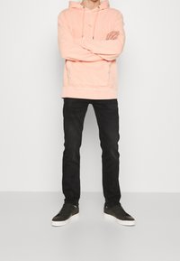 Osoba stojící se zkříženýma rukama, oblečená v světle růžové mikině s kapucí, černých slim džínech a černých teniskách s bílou podrážkou.