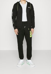 Persona che indossa una felpa nera con zip e pantaloni jogger con toppe logotipo bianche e verde neon, una maglietta bianca e sneakers bianche con dettagli verdi.