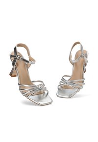 Sandalias de tacón alto metálicas plateadas con punta cuadrada, diseño de tiras cruzadas y un tacón bloque corto. Textura suave y hebilla ajustable.