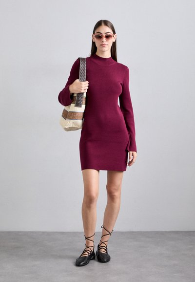 Nainen, jolla on päällään viininpunainen pitkähiha dress, ruskeat aurinkolasit, mustat nauhakengät ja joka kantaa vaaleanbeigeä ja ruskearaidallista olkalaukkua.