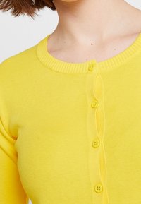 Yellow cardigan with a ribbed neckline, featuring five rounded buttons down the front. Fabric appears soft and slightly textured.