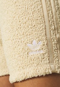 Light beige shorts made of textured fabric with a soft, fuzzy surface. Features gold side stripes and a white embroidered logo.