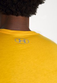 Žluté tričko s kulatým výstřihem, s jemnou texturou a šedým logem Under Armour tištěným blízko zadního límce.