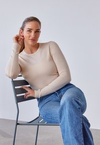 Beige langærmet top i blødt materiale med rund halsudskæring, kombineret med lyseblå brede jeans, siddende på en grå metalstol. Minimalistisk baggrund.