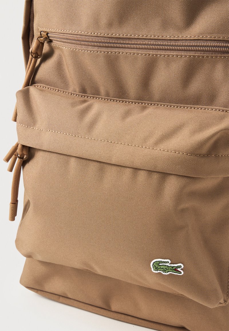 Zaino in tessuto beige, fronte con tasca con cerniera e toppa con logo coccodrillo verde nell'angolo in basso a destra.