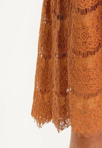 Blonde kjole i brændt orange med indviklede blomster mønstre, bølget kant og en semi-gennemsigtig tekstur.