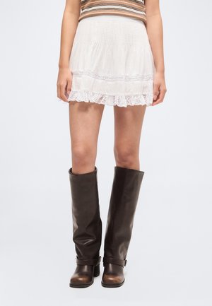 Személy fehér csipkés szegélyű miniszoknyában és barna, térdig érő bőr csizmában áll egyszerű háttér előtt.