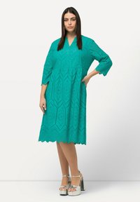 Turquoise jurk met geometrische uitsneden, driekwart mouwen en een uitlopende zoom. Draag met metallic plateauschoenen.