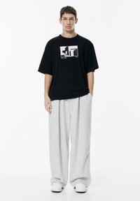 En ung mand står med front mod kameraet iført en sort grafisk t-shirt, løse lysegrå bukser og hvide sneakers mod en hvid baggrund.