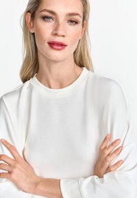 Felpa bianca con collo rotondo, maniche lunghe, texture liscia e vestibilità rilassata. Il modello ha i capelli biondo chiaro e un make-up neutro.