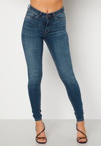 Vysoké skinny džíny v tmavě modrém denimu, které mají klasický pětikapsový design a jemné vyblednutí, doplněné černými sandály na podpatku.