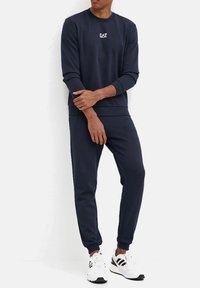 Felpa blu navy e pantaloni jogger in misto cotone. Presentano un piccolo logo bianco sul petto, polsini e caviglie a costine. Portati con sneakers bianche.