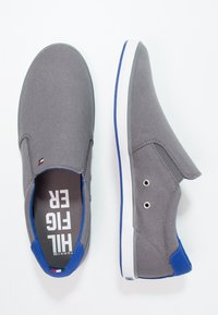 Scarpe slip-on grigie con accenti blu, tomaia in tessuto, suola in gomma bianca e occhielli in metallo. "HILFIGER" stampato sulle solette.