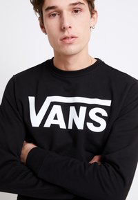 Czarna bluza z wyraźnym białym logo "VANS". Posiada okrągły dekolt i długie rękawy, a także gładki, teksturowany materiał.