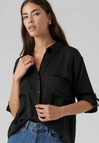 Camicia nera con bottoni realizzata in un tessuto leggero, con due tasche sul petto e maniche arrotolate. Il modello tiene il tessuto vicino alla vita.
