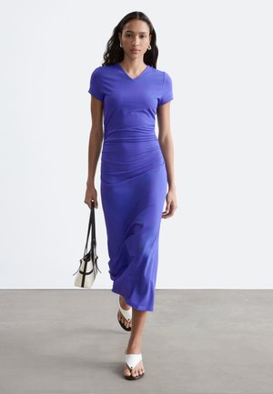 Femme marchant portant une robe midi violette ajustée et des sandales blanches, portant un sac à main noir et blanc avec de courtes poignées noires.