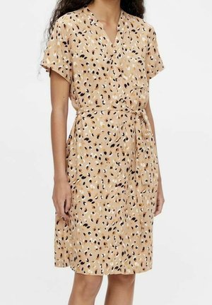 Mujer que lleva un vestido beige hasta la rodilla con manchas abstractas negras, blancas y marrones, mangas cortas y cinturón anudado en la cintura.