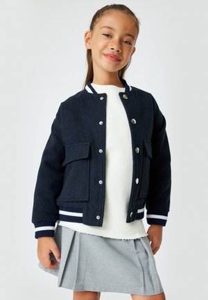 Giacca bomber navy con bottoni a pressione argentati, due tasche frontali, polsini a righe, indossata sopra un felpa bianca e una gonna plissettata grigia.