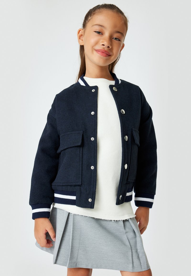 Chaqueta bomber azul marino con botones a presión plateados, dos bolsillos delanteros, puños de canalé a rayas, que se lleva sobre un sudadera blanca y una falda gris con pliegues.