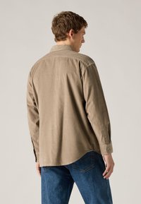 Καφέ πουκάμισο με μακριά μανίκια, γιακά, ευθεία γραμμή στο τελείωμα και μανσέτες με κουμπιά. Φοριέται με μπλε τζιν, παρουσιάζοντας ένα casual, χαλαρό στυλ.