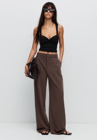 Černý přiléhavý crop top s otočeným detailem, zkombinovaný s volnými hnědými kalhotami s širokými nohavicemi. Modelka drží malou tmavou kabelku a nosí sluneční brýle.