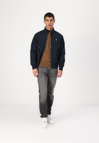 Giubbotto bomber blu navy con colletto a coste, due tasche laterali e logo giallo, abbinato a un maglione marrone, jeans grigi e sneakers bianche.