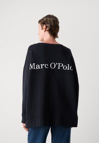 Didelis juodas megztinis su baltu "Marc O'Polo" logotipu ant nugaros. Minkšta medžiaga, laisvas fasonas ir gumuoti rankogaliai.