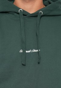 Hoodie vert en tissu doux, doté d'une poche kangourou, de cordons de serrage réglables et de texte brodé blanc sur la poitrine.