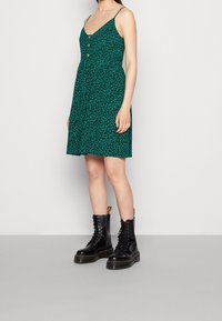 Vestido verde con un estampado floral, escote en V y detalle de botones, combinado con botas negras de cordones. Presenta tirantes finos y una cintura fruncida.