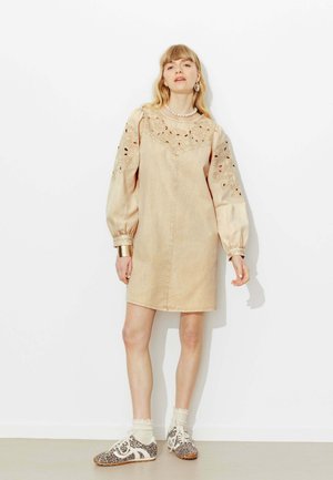 Une femme se tient debout, portant une robe beige avec des manches bouffantes brodées, un collier de perles, un bracelet manchette et des chaussures imprimé léopard avec des chaussettes blanches en dentelle.