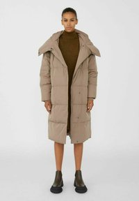 Manteau rembourré beige long avec un col montant, doté d'une fermeture éclair à l'avant et de boutons-pression. Stylisé sur une robe tricot brun, associé à des bottes noires à semelles épaisses.